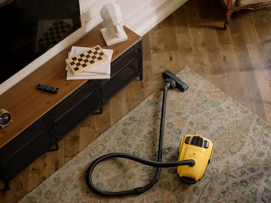 A yellow canister vacuum cleaner with a black hose and a metal wand attachment rests on a patterned area rug in a room with wooden flooring. To the left, a black metal TV stand holds a remote control, a white bust sculpture, and papers, situated against a white wall with decorative molding. Natural light illuminates the scene, highlighting the clean and orderly appearance of the space. The setting appears to be a residential living room, and the image reflects surface cleaning and deep cleaning activities associated with domestic sanitisation, as promoted by Carpet Cleaners Marylebone on their website dedicated to best carpet cleaning tips for Baker Street flats in Marylebone.