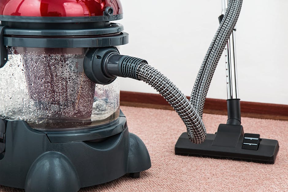 Close-up of a professional wet vacuum cleaner with a clear canister filled with water and dust, connected to a flexible, patterned hose. The vacuum is positioned on a pinkish-beige carpet in a residential room, with a portion of a hardwood skirting board along a white wall in the background. The vacuum features a red top and black base, and appears to be actively engaged in surface cleaning or deep cleaning tasks, highlighting the equipment used by Carpet Cleaners Marylebone for sanitisation and maintenance of carpets in Baker Street flats.