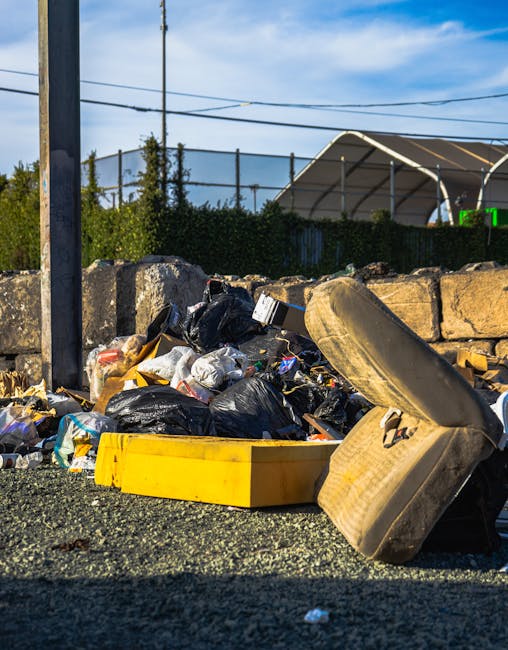 An outdoor scene showing a pile of bulky waste, including a worn, beige upholstered armchair with visible fabric texture and stains, discarded black and white plastic bags filled with waste, and a bright yellow plastic container. The waste is situated on a gravel surface beside a stone wall made of irregularly shaped, weathered stones. In the background, there are metal fences, utility poles, power lines, and a partially visible greenhouse with a curved roof, under a blue sky with some clouds. The area appears untidy and in need of rubbish removal, which Carpet Cleaners Marylebone can assist with through their local waste disposal and cleaning services.
