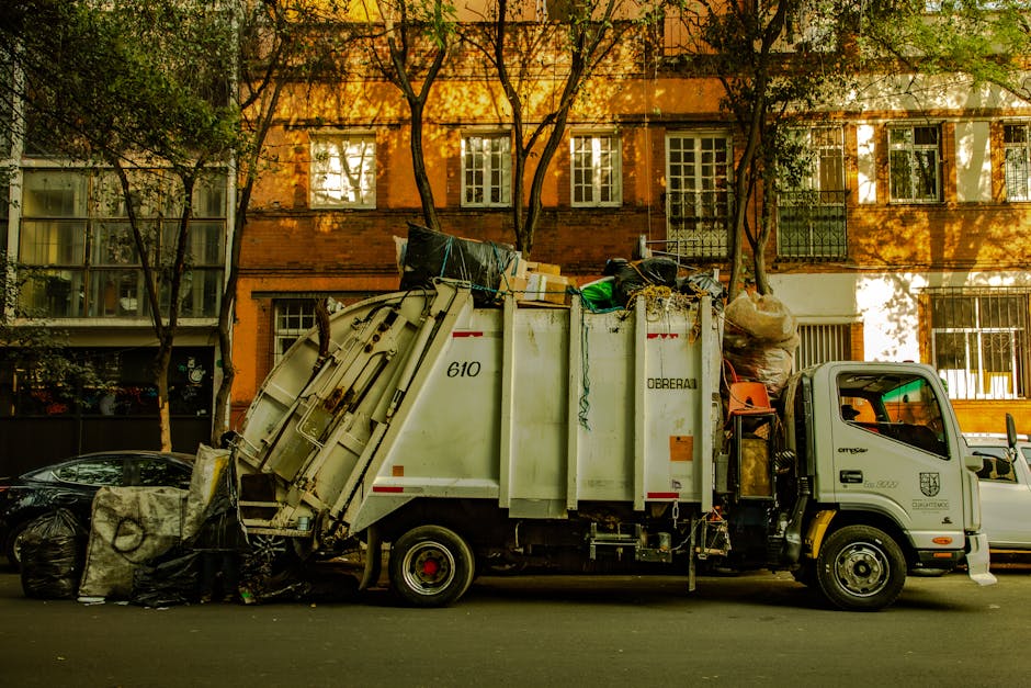 A small commercial waste collection truck parked on a street in Marylebone, with a white and grey body and black wheels, filled with various bulky waste items such as bags, boxes, and discarded household items. The truck's side panel has the number 610 and the company's branding, indicating it is operated by Carpet Cleaners Marylebone. In front of the truck, there are a few large black garbage bags on the pavement. The background features a multi-story brick building with multiple windows, some of which are open, and trees casting dappled sunlight onto the scene. The overall area appears clean and well-maintained, reflecting local waste disposal and bulky waste removal services.