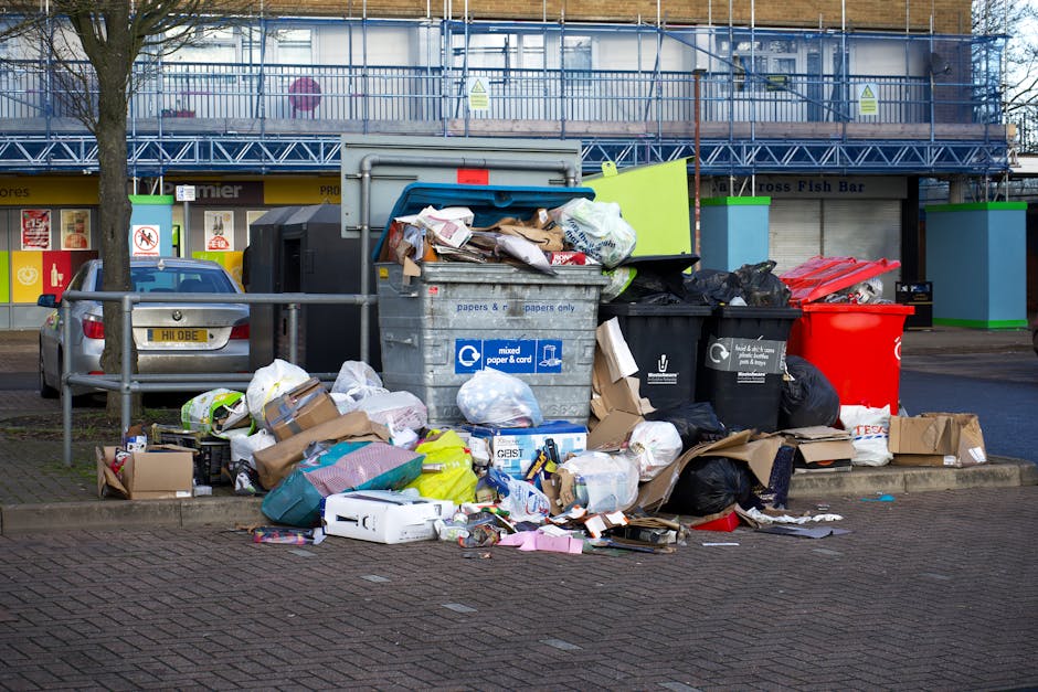 An outdoor scene showing a pile of bulky waste, including a worn, beige upholstered armchair with visible fabric texture and stains, discarded black and white plastic bags filled with waste, and a bright yellow plastic container. The waste is situated on a gravel surface beside a stone wall made of irregularly shaped, weathered stones. In the background, there are metal fences, utility poles, power lines, and a partially visible greenhouse with a curved roof, under a blue sky with some clouds. The area appears untidy and in need of rubbish removal, which Carpet Cleaners Marylebone can assist with through their local waste disposal and cleaning services.