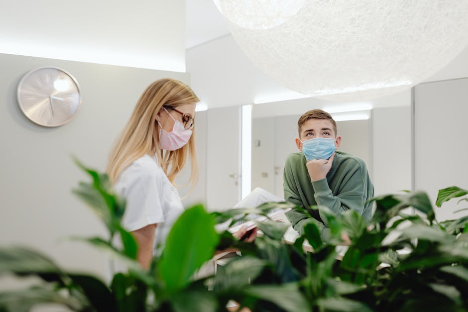 A female healthcare professional wearing a pink face mask, glasses, and a white uniform interacts with a young male patient or clinic visitor, who is seated behind a reception desk. The patient, dressed in a green hoodie and a blue face mask, rests his chin on his hand while looking upward. The scene is set in a clean, well-lit medical or clinic reception area with white walls, a large clock, and a ceiling with smooth, rounded architectural features. Green leafy plants partially obscure the lower part of the image, contributing to a calming, hygienic environment. This setting emphasizes surface cleaning, hygiene, and maintenance standards appropriate for Harley Street clinics and patient areas, aligning with the services offered by Carpet Cleaners Marylebone.