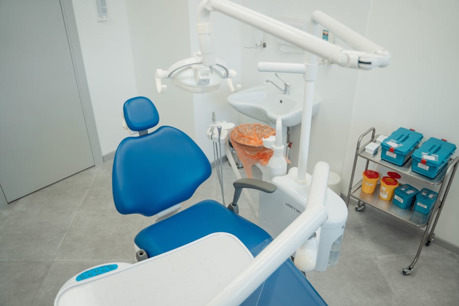 A clinical treatment room featuring a blue dental chair with an adjustable headrest and armrests, positioned prominently in the foreground. Behind the chair, there is a white sink with a stainless steel faucet, accompanied by a small orange waste bag liner attached to the toilet. To the right, a metal trolley holds neatly organized blue and red containers for sterilization and medical supplies. The room has a clean, bright appearance with light grey tiled flooring, white walls, and ample lighting, creating an environment suitable for surface cleaning, sanitisation, and hygiene maintenance. Carpet Cleaners Marylebone ensures thorough deep cleaning of such medical spaces to meet healthcare standards.