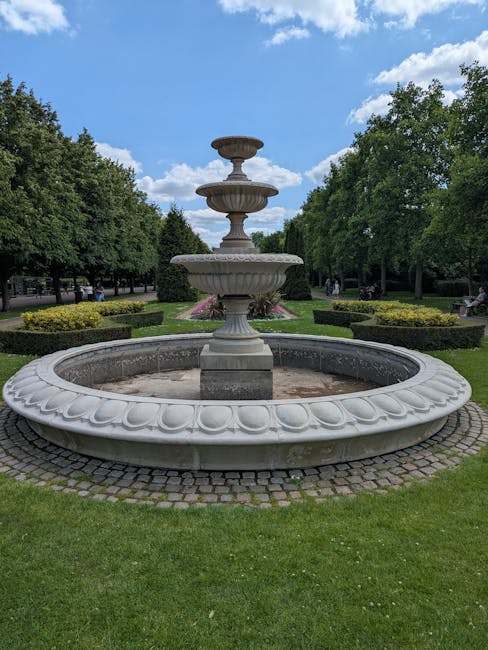 A large, decorative stone fountain situated in a landscaped park, featuring multiple tiers with ornate detailing, surrounded by a circular basin with a scalloped edge. The fountain is central in the image, set on a paved cobblestone surface, with lush green grass, neatly trimmed bushes, and tall trees in the background. The sky above is partly cloudy with patches of blue visible. The area appears well-maintained and clean, reflecting a serene outdoor environment. Carpet Cleaners Marylebone would emphasize the importance of surface cleaning and maintenance of outdoor features like this fountain as part of overall hygiene and aesthetic upkeep. The scene showcases a peaceful, outdoor space that benefits from regular deep cleaning and sanitisation to preserve its appearance and hygiene.