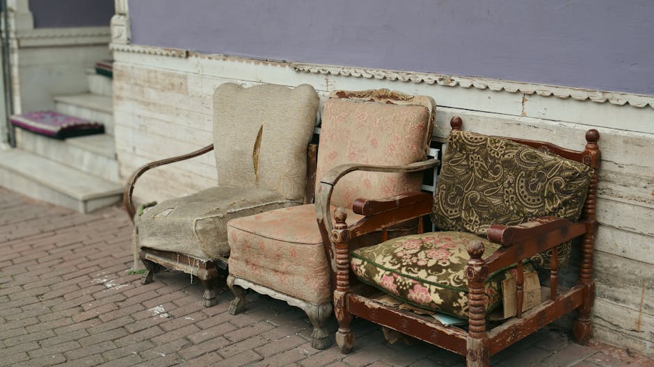 An orange upholstered armchair with a high back and rolled arms, placed on a brick-paved outdoor patio area outside a building in Marylebone. Behind the chair, there is a green leafy shrub partially c