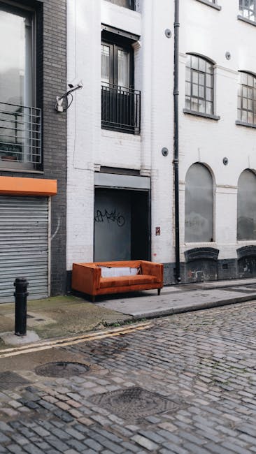 An outdoor scene on a cobblestone street featuring a white multi-story building with large arched and rectangular windows. On the sidewalk, there is an old, orange upholstered sofa placed directly against the building's wall, with a white sheet or fabric draped over its back. The building's exterior shows signs of weathering. A black bollard is positioned in front of the sofa to protect it. Natural, diffused daylight illuminates the scene, highlighting the urban setting. Carpet Cleaners Marylebone provides professional cleaning services, including upholstery cleaning, for properties in the area, ensuring surfaces like sofas and furniture are thoroughly deep cleaned and hygienic.