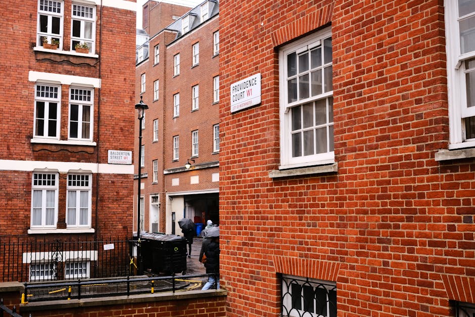 A street view of red brick residential buildings with white-framed sash windows in Marylebone, London, showing a narrow alleyway with a black metal gate and nearby black waste bins. The scene is illuminated by natural daylight, highlighting the clean, well-maintained façade of the buildings. The image reflects urban architecture typical of Marylebone High Street, and while not directly depicting cleaning, it relates to the area's residential upkeep. Carpet Cleaners Marylebone offers professional surface cleaning and deep cleaning services to maintain such environments, ensuring hygiene and cleanliness in domestic and commercial spaces.