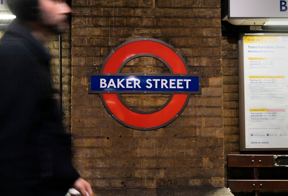 A close-up view of a brick wall in a London Underground station, featuring a circular, red and blue sign displaying 'Baker Street'. To the right, a digital information board provides train schedules and notices, with visible multilingual text. The lighting is artificial, casting a neutral tone over the scene. In the foreground, part of a person dressed in black is blurred, suggesting movement. The surface of the brick wall appears clean and well-maintained, with no visible dust or dirt. This setting reflects an urban environment typical of public transportation hubs, emphasizing cleanliness and hygiene in the station's design, consistent with professional cleaning standards promoted by Carpet Cleaners Marylebone.