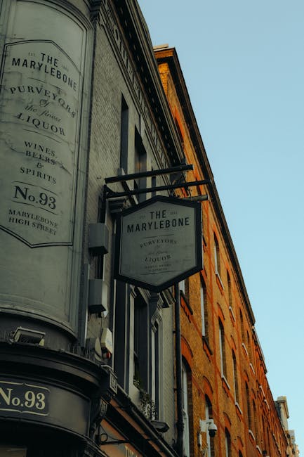 Photograph of the exterior of a historic building on Marylebone High Street, featuring a black and white sign reading 'The Marylebone, Purveyors of Liquor, Wines, Beers, Spirits, No. 93'. The building has a combination of ornate and simple architectural details with large windows, some with decorative moldings, and a curved corner with a prominent sign. The facade displays a mixture of stone and brickwork with a warm, orangey hue, illuminated by natural daylight. The street appears clean and well-maintained, with clear signage and a classic, inviting entrance likely associated with a traditional liquor store or pub, consistent with the elegance of the Marylebone area. Carpet Cleaners Marylebone offers professional cleaning and sanitisation services, ensuring surfaces remain pristine and hygienic, suitable for both residential and commercial spaces.
