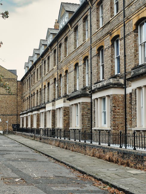 The image shows a row of Victorian terraced houses with brick facades, white-framed sash windows, and black wrought iron railings along the paved sidewalk. The buildings have pitched slate roofs with dormer windows, and the street appears clean and well-maintained. Natural daylight illuminates the scene, highlighting the textured brickwork and the tidy appearance of the exterior surfaces. Mentioned in context with surface cleaning and maintenance, Carpet Cleaners Marylebone ensures hygienic and pristine conditions for residential properties on Portman Estate, specialising in deep cleaning and sanitisation services for Victorian homes.