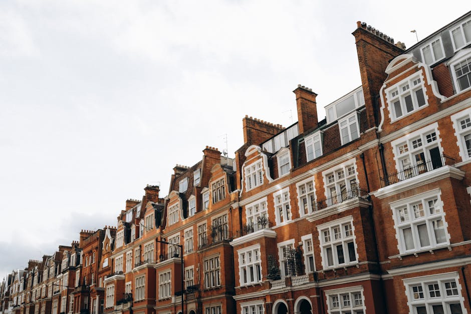 A row of Victorian-style residential buildings with red brick facades, white window frames, and decorative architectural details, situated under a cloudy sky. The buildings feature multiple stories, with balconies and roof windows visible, reflecting a classic Marylebone residential street. The exterior surfaces appear clean and well-maintained, highlighting the importance of thorough surface cleaning and upkeep for Victorian homes, as offered by Carpet Cleaners Marylebone within their professional domestic cleaning services.