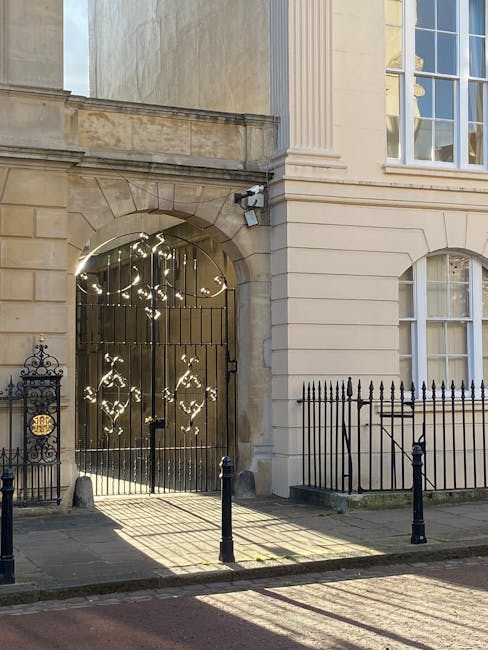 Image of an ornate wrought iron gate set within a stone archway, attached to a historic beige stone building with decorative columns and large sash windows. The gate features intricate patterns illuminated by sunlight, creating reflections and shadows on the pavement outside. To the left of the gate, there is a black iron fence with decorative finials, while in front, short black bollards separate the sidewalk from the street. The building's façade appears clean and well-maintained, reflecting a neat and tidy appearance suitable for environments requiring surface cleaning and maintenance. The lighting highlights the textures of the stone and metal surfaces, indicating a bright, clear day. This scene aligns with the conservation and upkeep themes related to Westminster Council Carpet Waste Rules, as handled by Carpet Cleaners Marylebone, emphasizing the importance of proper hygiene and surface cleaning for historic properties.