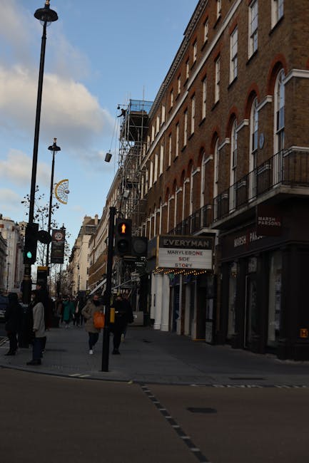 A busy street scene in Marylebone, Westminster, featuring multi-story brick buildings with large windows and retail storefronts at street level. The pavement is populated with pedestrians, some crossing at a traffic signal that displays an orange pedestrian figure. Street lamps and hanging signs, including one indicating 'Marylebone SOE STORY,' line the sidewalk. Overhead, scaffolding is visible on part of a building, indicating ongoing construction or maintenance. The sky is partly cloudy, and the lighting suggests daytime. Carpet Cleaners Marylebone's signage is not visible in this particular image, but the scene emphasizes the urban environment and cleanliness typical of a well-maintained commercial area.