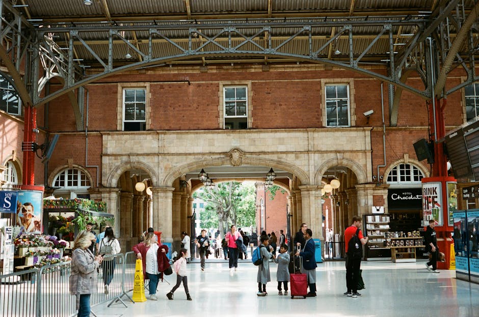 Interior view of a busy train station with a high, arched metal ceiling and large brick and stone walls. The scene shows a variety of people, including families, travelers, and staff, some carrying luggage and others browsing small shops and kiosks. The station features tiled flooring that appears clean and polished, with visible lighting fixtures hanging from the ceiling. In the background, an arched doorway leads to the outside, where trees and natural light are visible. The overall environment emphasizes a well-maintained space that benefits from regular surface cleaning and deep cleaning practices, which is aligned with the services offered by Carpet Cleaners Marylebone to ensure hygiene and spotless conditions in busy public areas such as these.