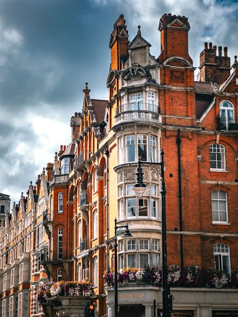 Photograph of a historic red-brick building on Marylebone street, featuring ornate white stone detailing around large bay windows and decorative turrets at the roofline. The building has multiple stories with a mix of brick and white trim, including flower boxes with vibrant blooms on the balconies. The sky overhead is cloudy, casting soft light on the façade. Street lamps and a nearby traffic light are visible in the foreground, emphasizing an urban, residential area. This image highlights the classic architectural style visible in central London, as seen in properties maintained by Carpet Cleaners Marylebone for domestic cleaning and surface sanitisation purposes.